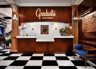 he Graduate Hotel New Haven lobby featuring a vintage-style front desk with wood paneling, black-and-white checkered floors, a wall of antique keys, and a cozy blue velvet bench.
